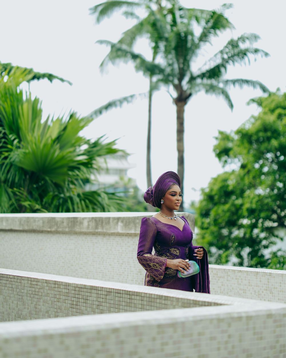 A person in a purple dress poses thoughtfully against a curved white wall with tropical palm trees in the background.