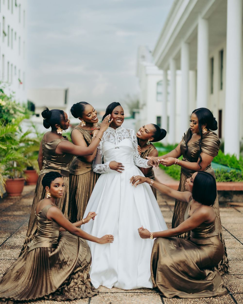 Wedding party in brown bridesmaid dresses playfully pose with bride in white gown against white columns outdoors.