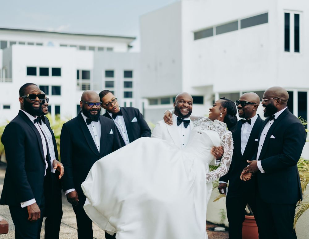 Wedding guests in formal black suits and tuxedos gather outdoors for a celebration.