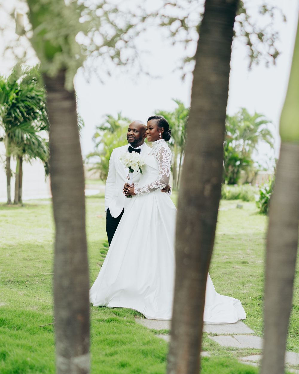 A couple in wedding attire shares an intimate moment among palm trees in a lush outdoor garden setting.