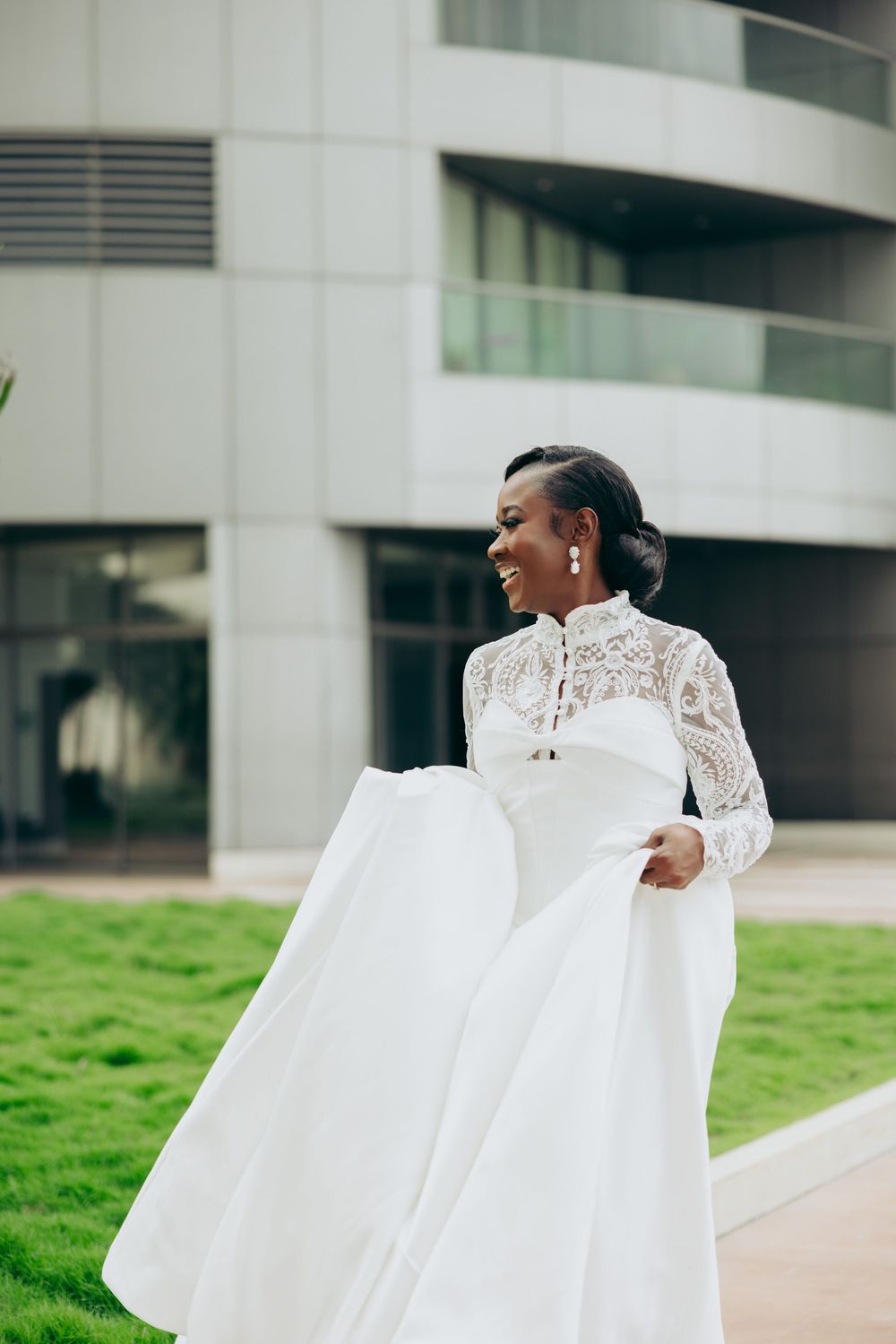 A bride in a flowing white lace wedding dress twirls joyfully outside a modern building.