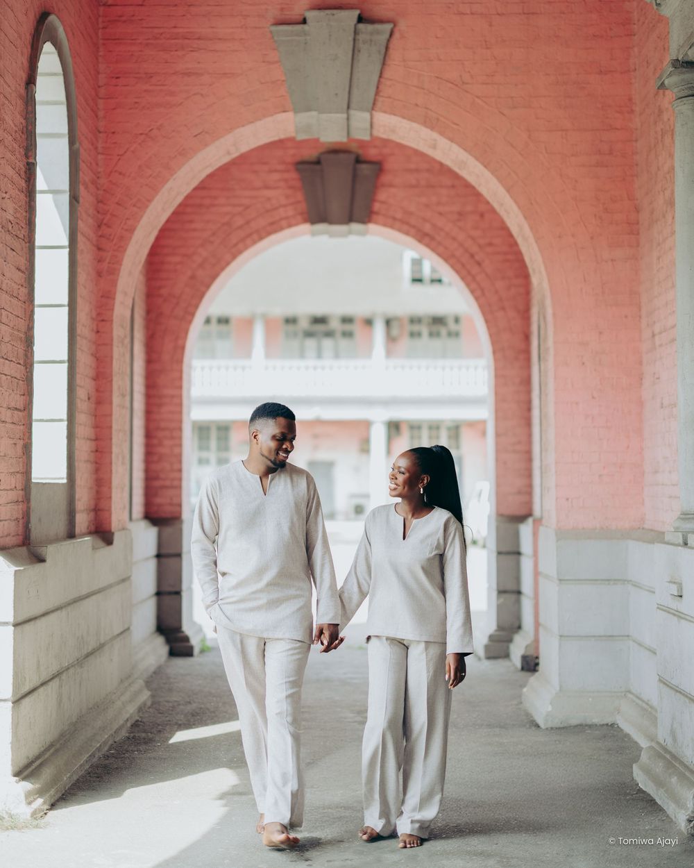 Couple in white outfits walking through a pink-walled colonnade with arched architecture and natural lighting.