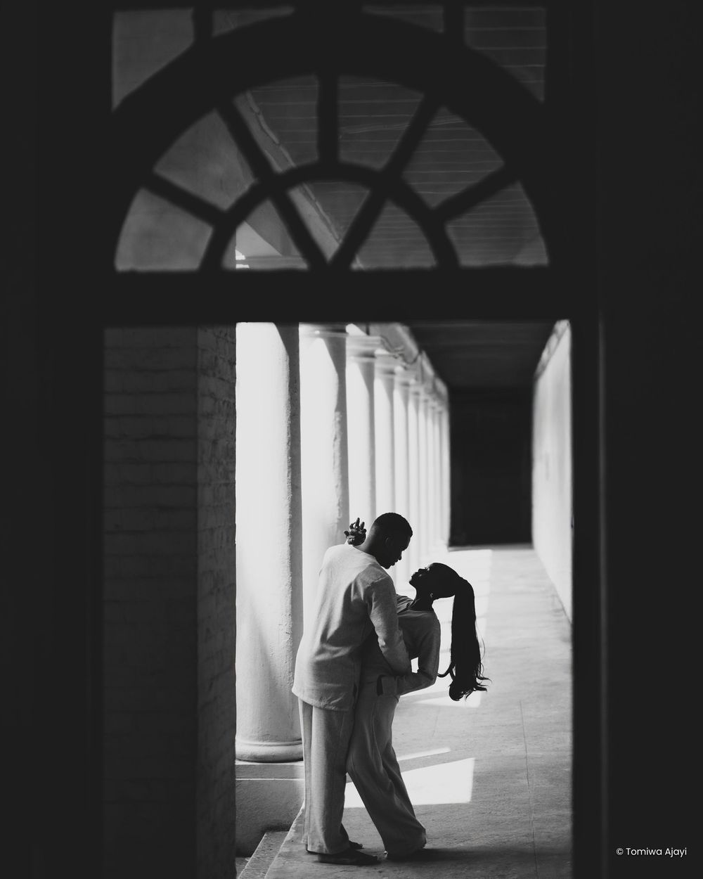 A dramatic black and white architectural photograph of a colonnade hallway with arched windows creating repeating shadows.