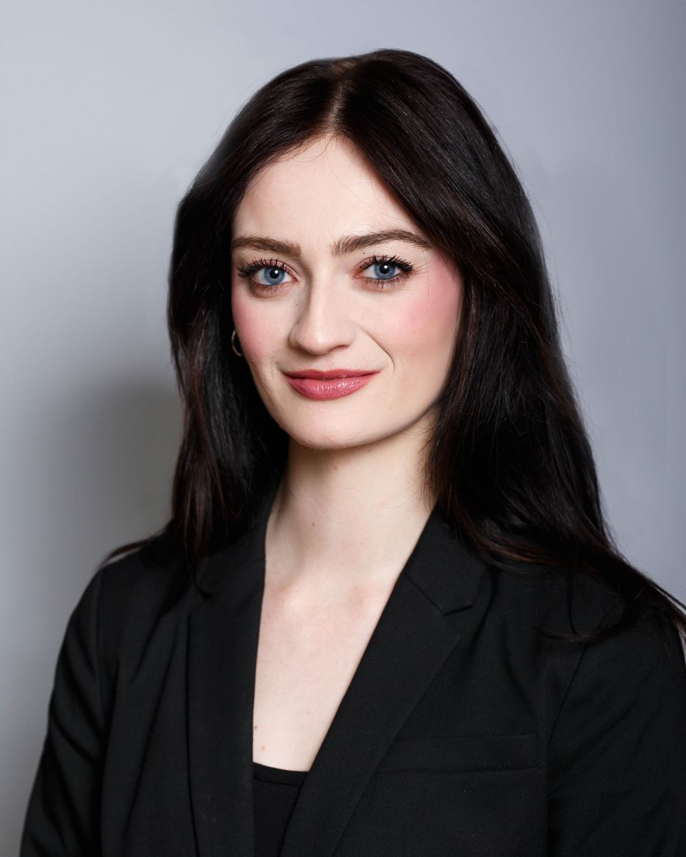 Professional headshot portrait of a person with dark hair wearing a black blazer against a light gray background.