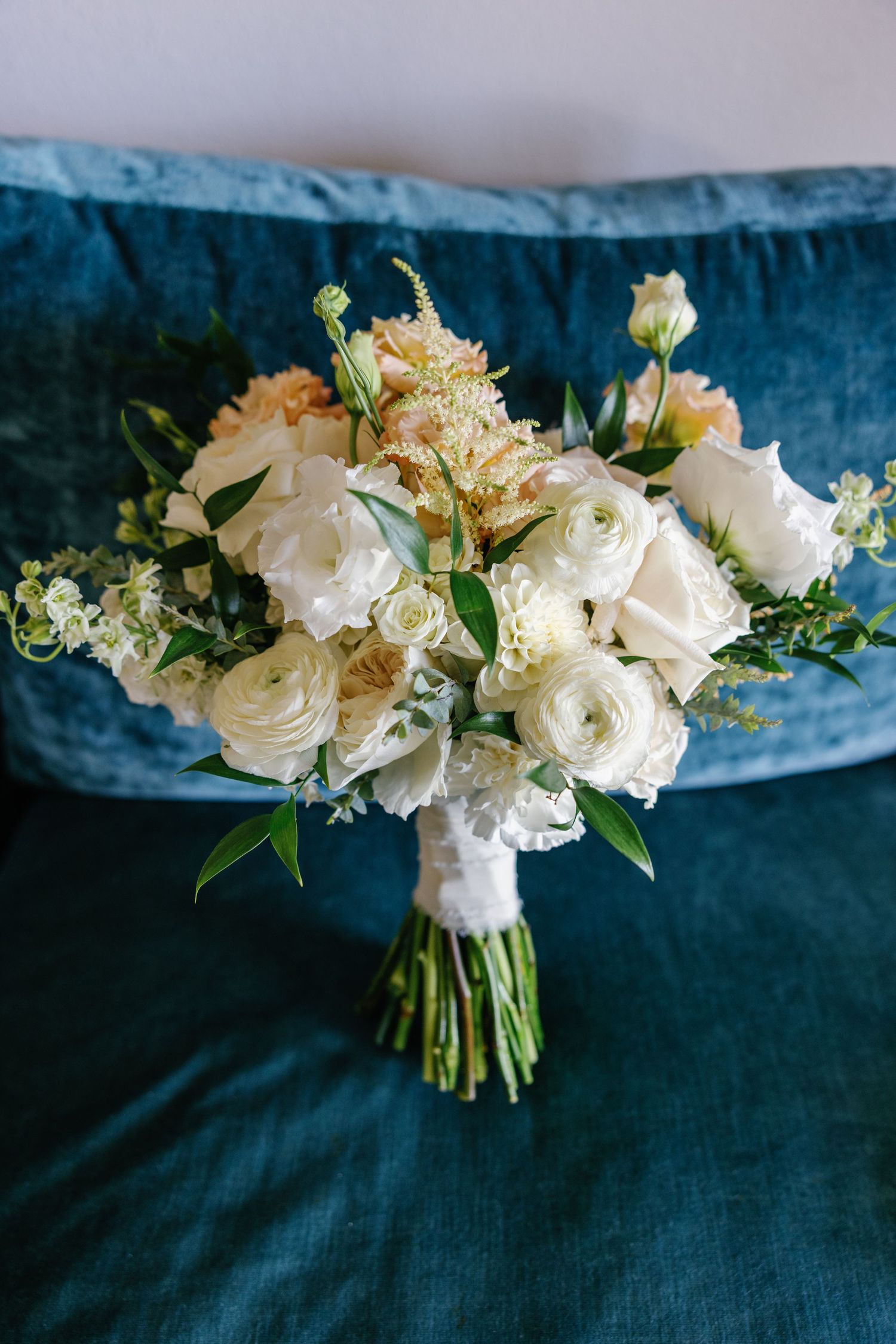 Elegant bridal bouquet of white roses and ranunculus resting on teal velvet sofa.