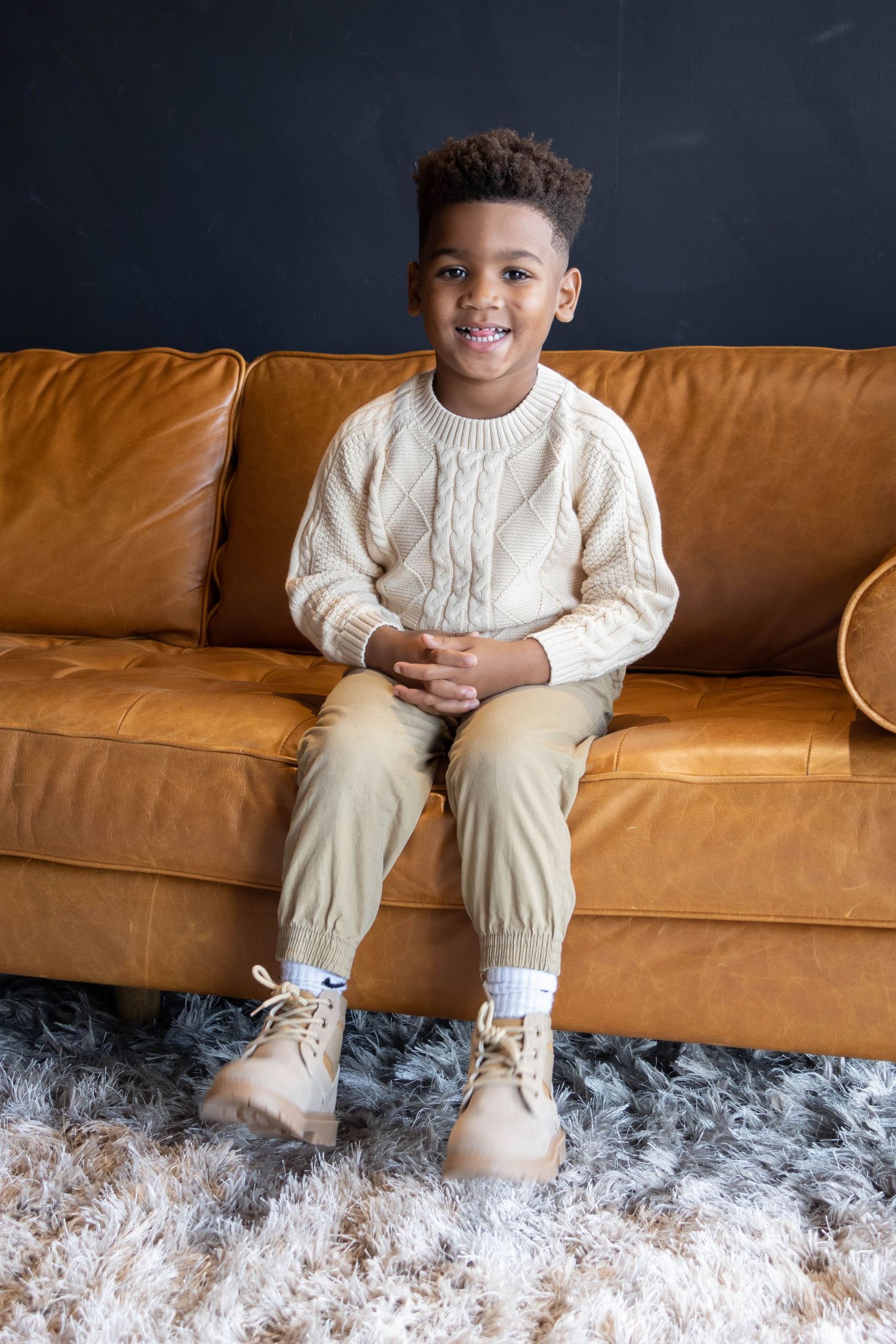 African American boy on a tan couch smiling a silly smile wearing tan and cream.Salem and Gloucester County Children Photography