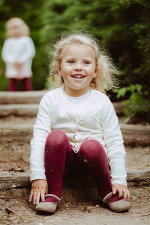 Child in white sweater and burgundy pants sitting on ground among trees.