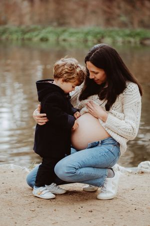 A tender moment between a pregnant woman in a white sweater and child by a lake.