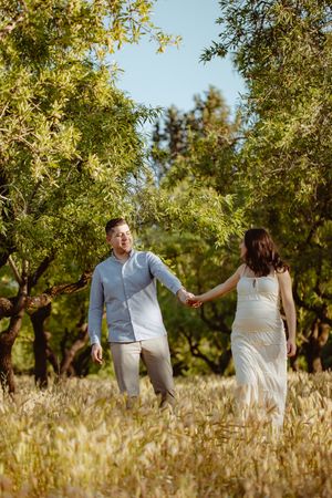 A couple walks hand in hand through a sunlit field with golden grass and trees in the background.
