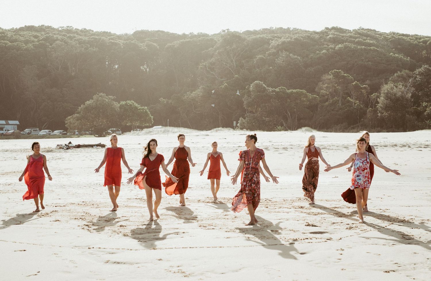 People in flowing red dresses running together along a sandy beach.