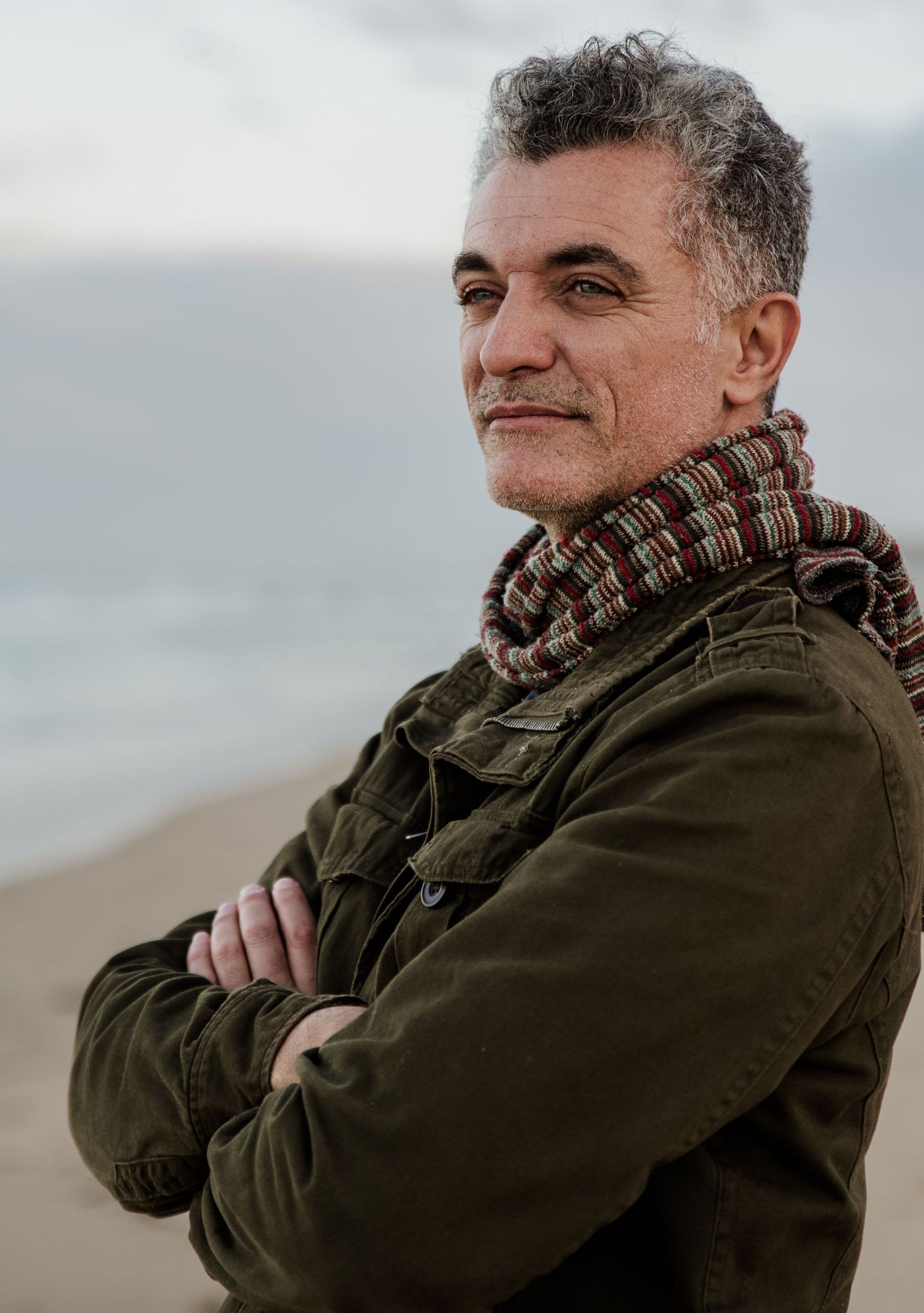 Portrait of someone in an olive jacket and patterned scarf at the beach.
