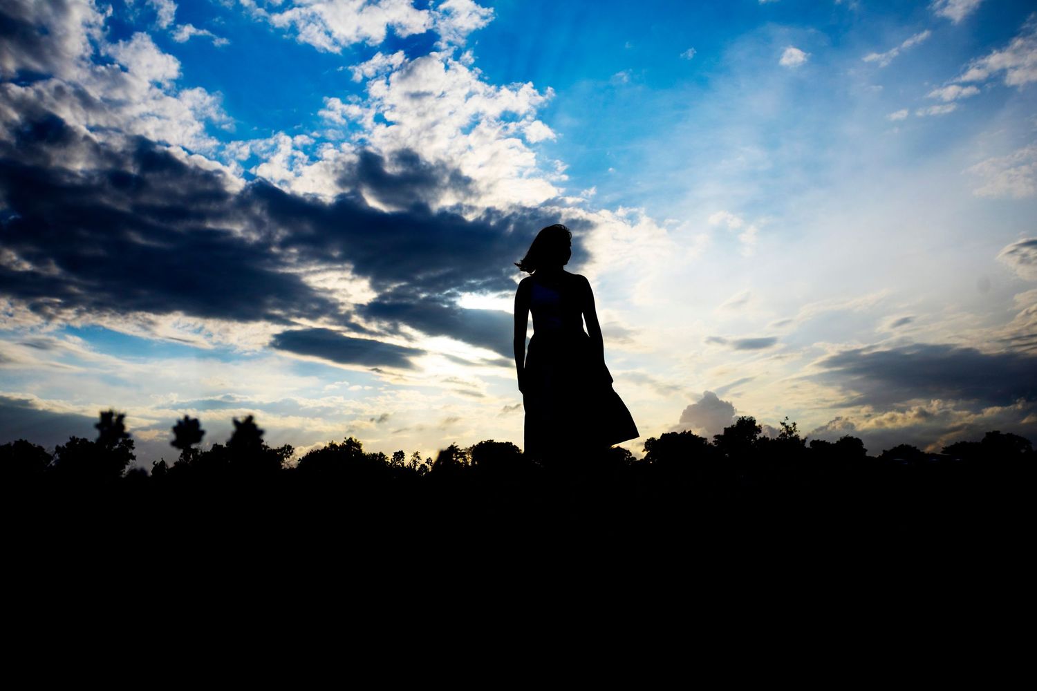 Silhouette of a woman in a dress and hat standing against a dramatic blue sky with clouds at sunset.
