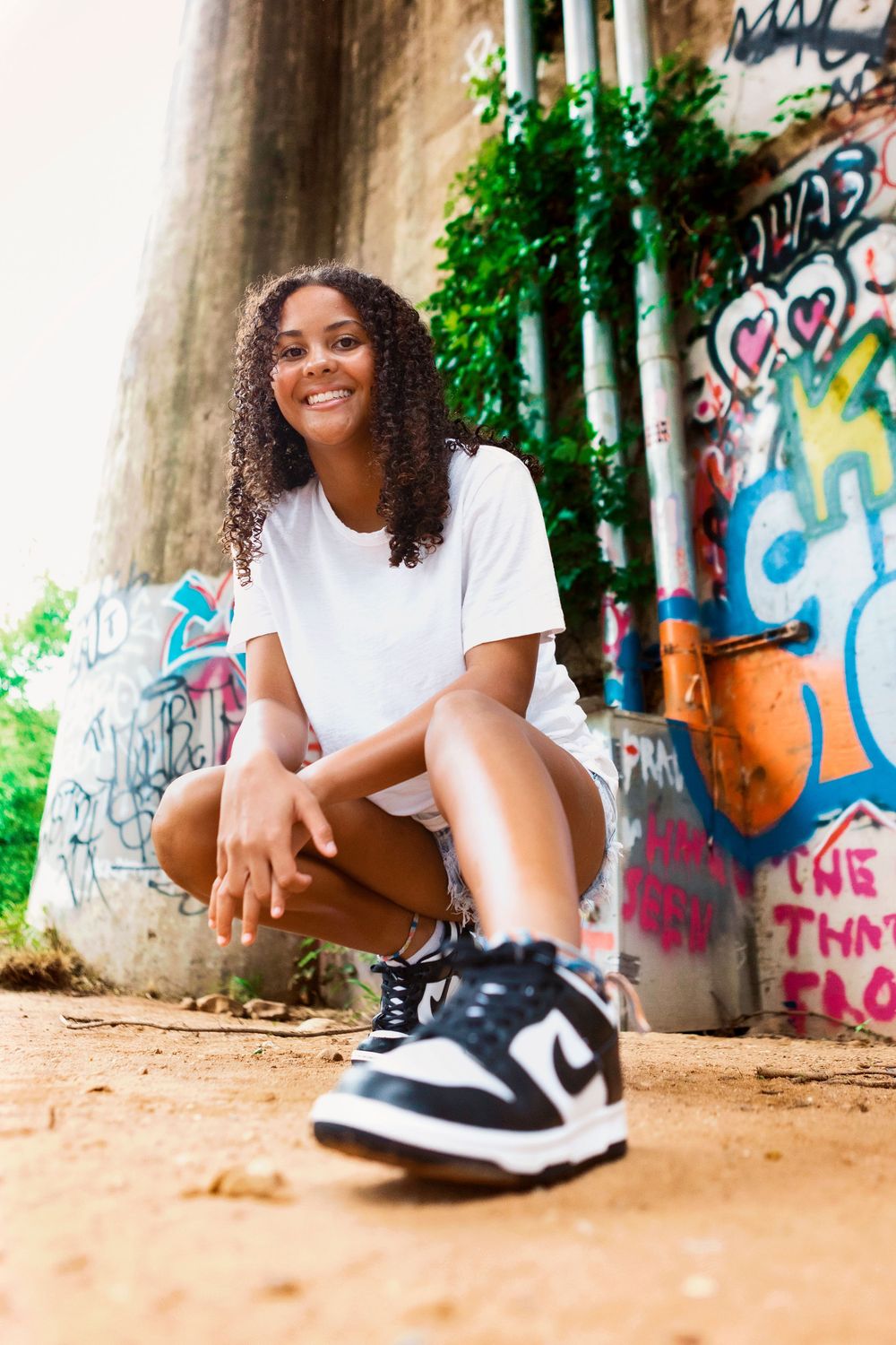 Portrait by graffiti wall featuring person in white t-shirt, jean shorts and Nike sneakers sitting casually outdoors.