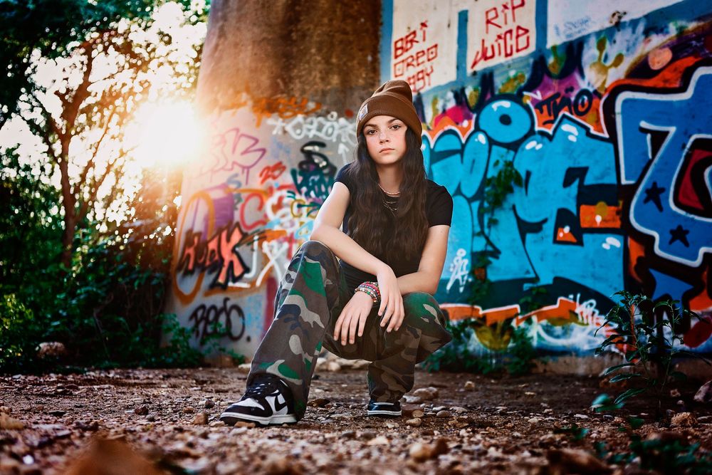 A person sits against a graffiti wall at sunset wearing casual attire with colorful street art in the background.