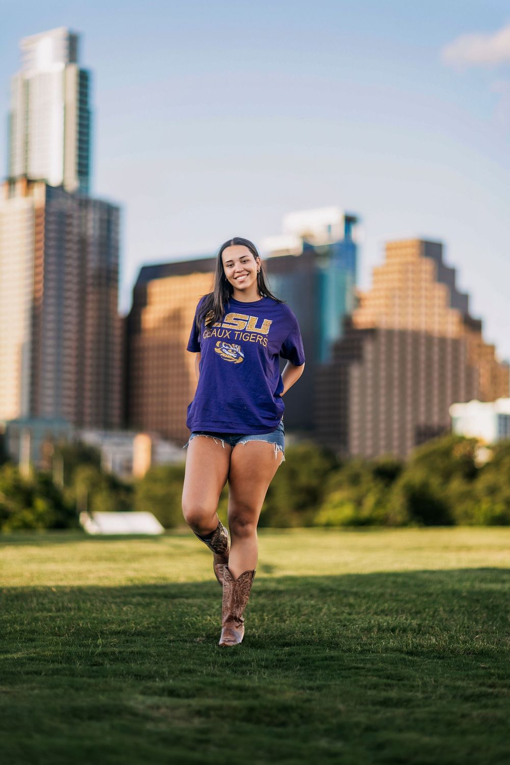 Young woman smiling in LSU Tigers purple shirt and denim shorts with cowboy boots on green lawn with city skyline.