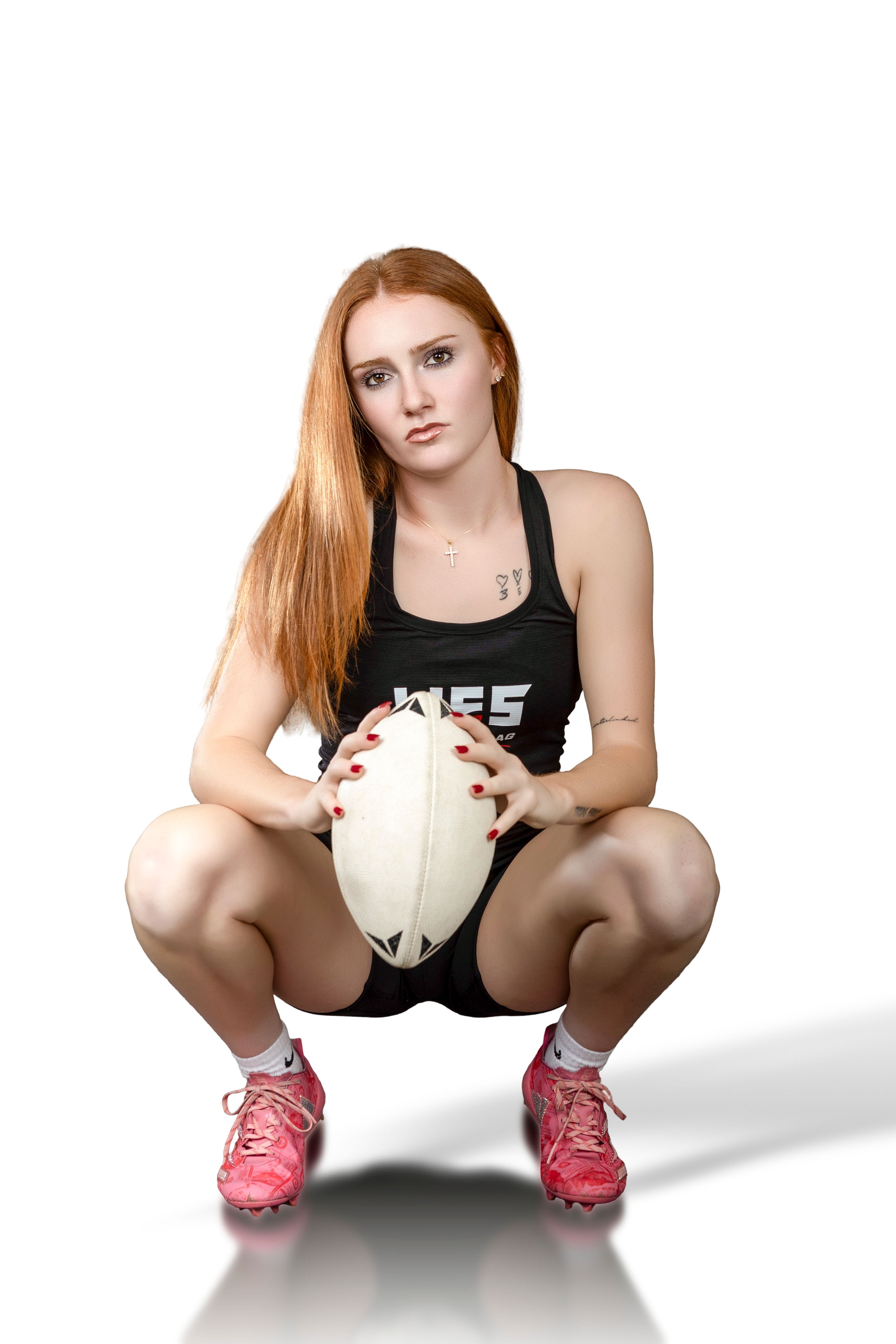 Female rugby player with red hair crouching in black uniform, holding a rugby ball, wearing pink cleats on white background.