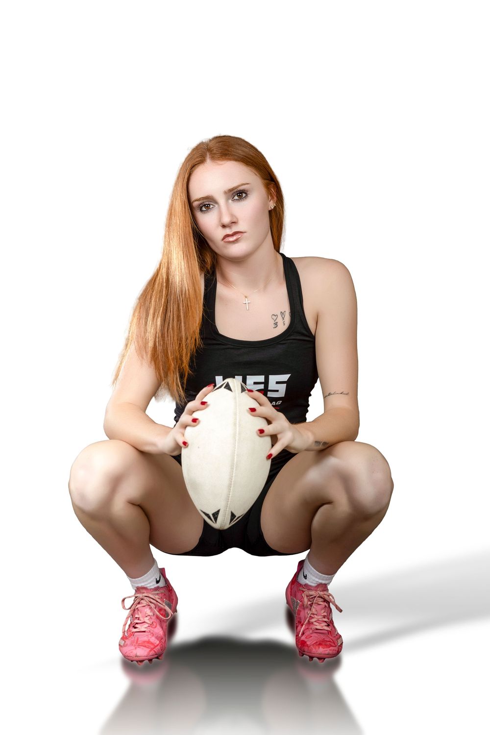 Female rugby player with red hair crouching in black uniform, holding a rugby ball, wearing pink cleats on white background.