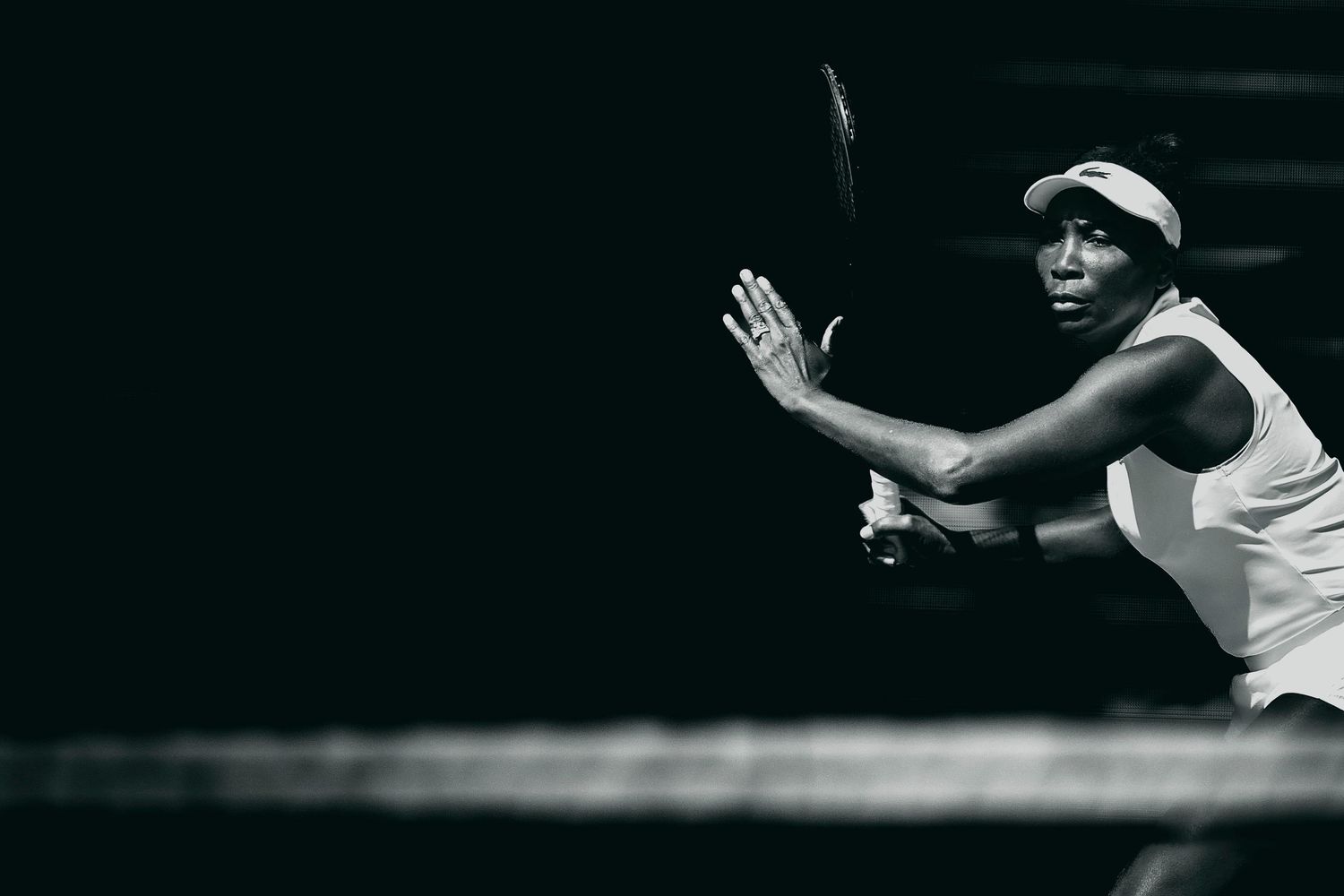 Black and white photo of a female tennis player mid-swing, wearing a white outfit and cap on a dark court.