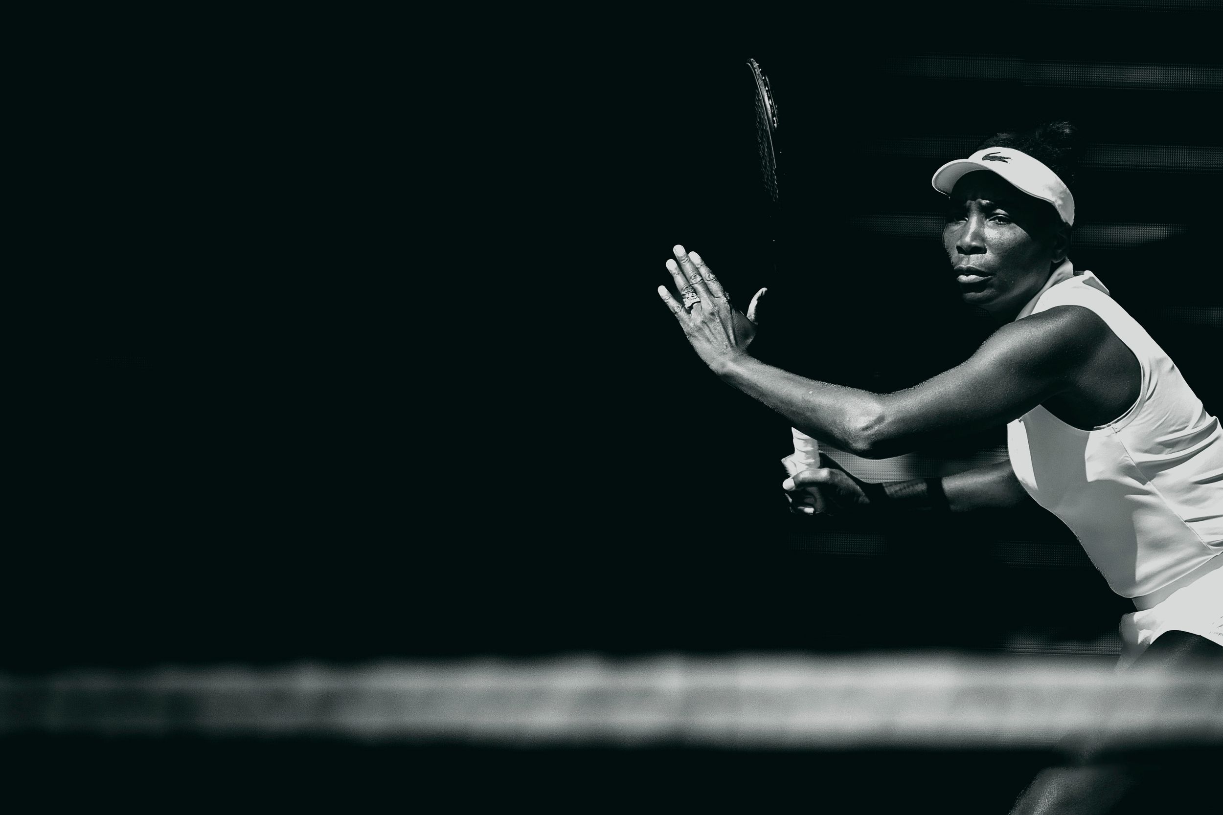 Black and white photo of a female tennis player mid-swing, wearing a white outfit and cap on a dark court.