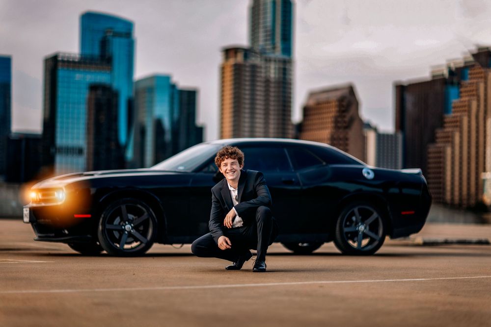 Young man in suit poses crouching before black Dodge Challenger on rooftop parking with city skyline at dusk.