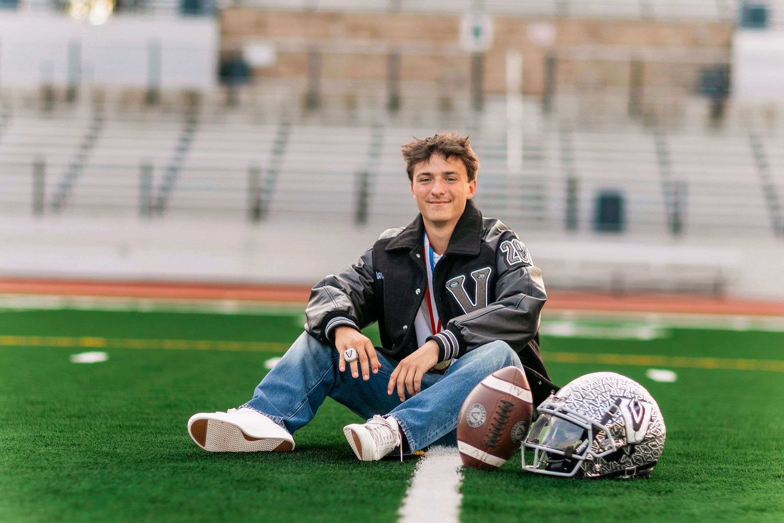Teen boy in varsity jacket sits on football field with helmet and ball for senior portrait photo.