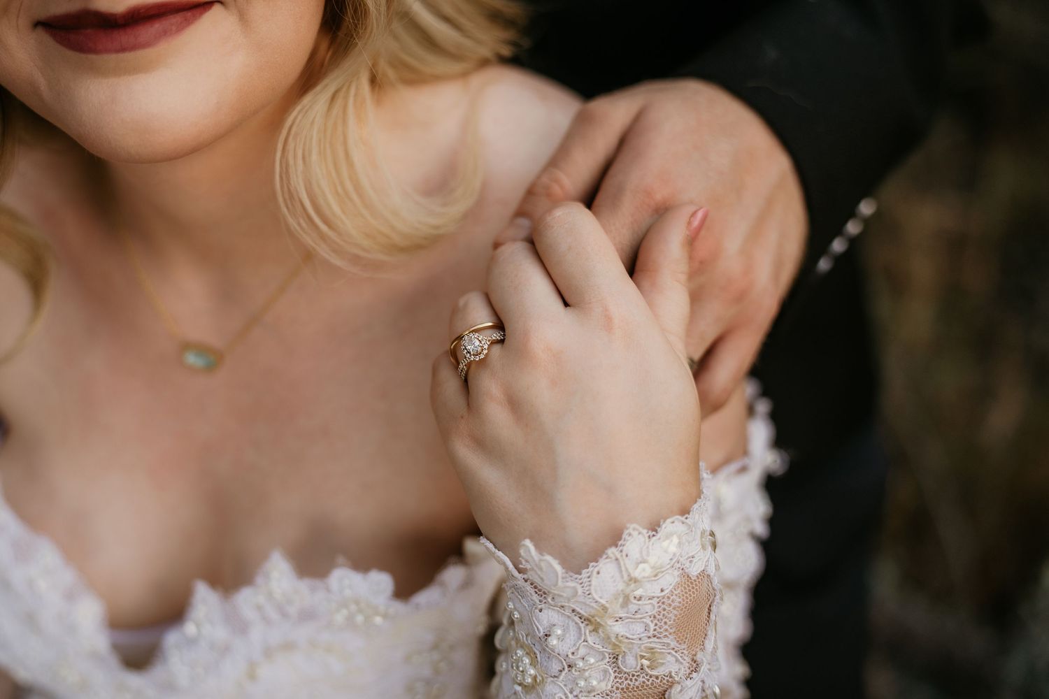 Close-up of bride wearing lace wedding dress and diamond ring, with groom's hand resting on her shoulder.