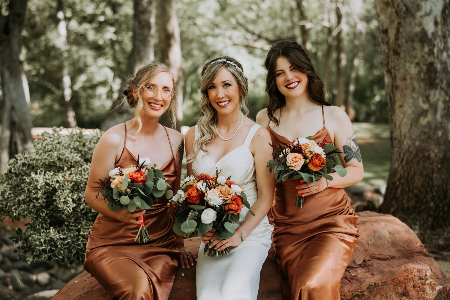 Three women in earth-toned dresses pose with floral bouquets in a garden setting.