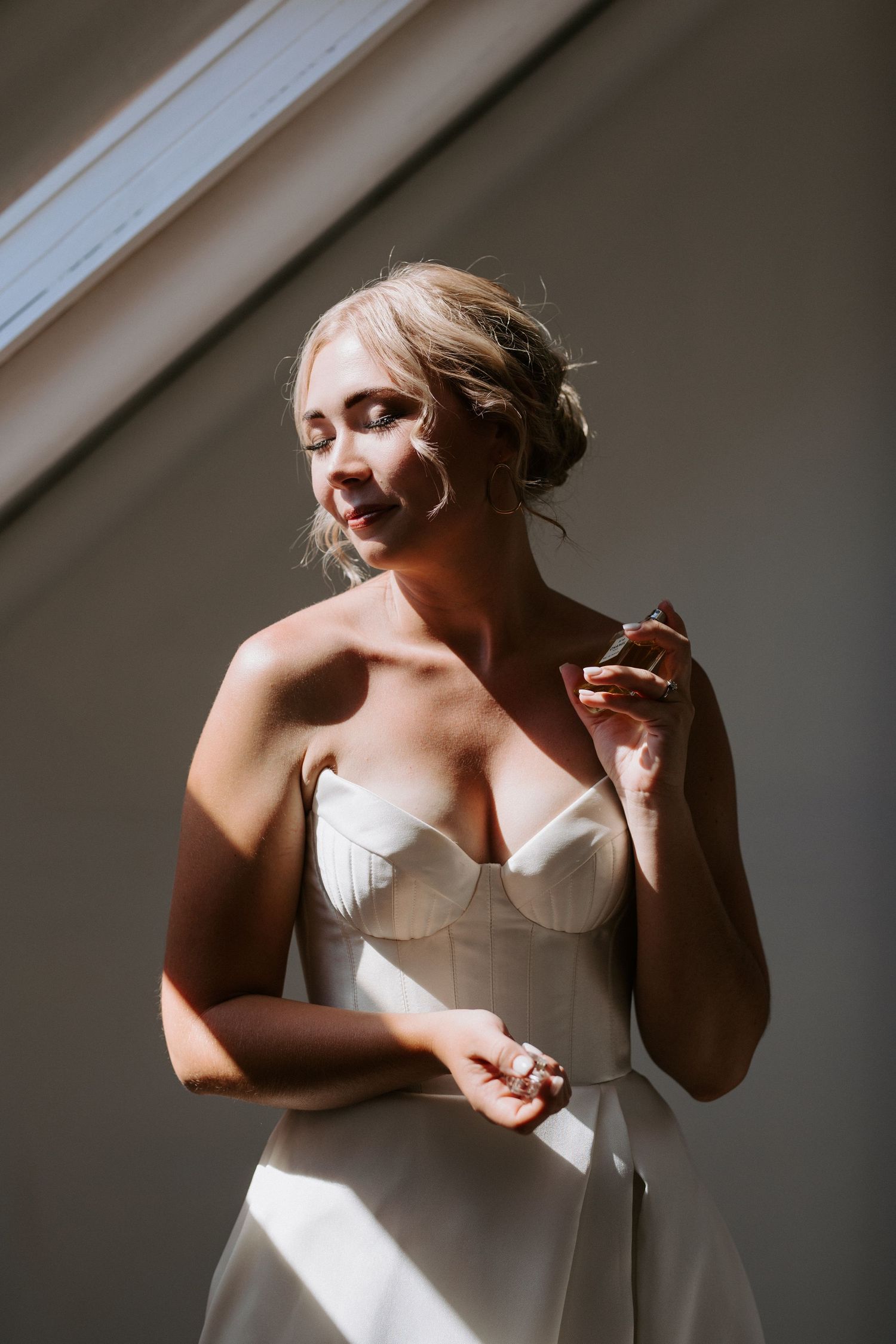A bride stands in front of a sunlit window, eyes closed as she delicately sprays perfume before her ceremony.