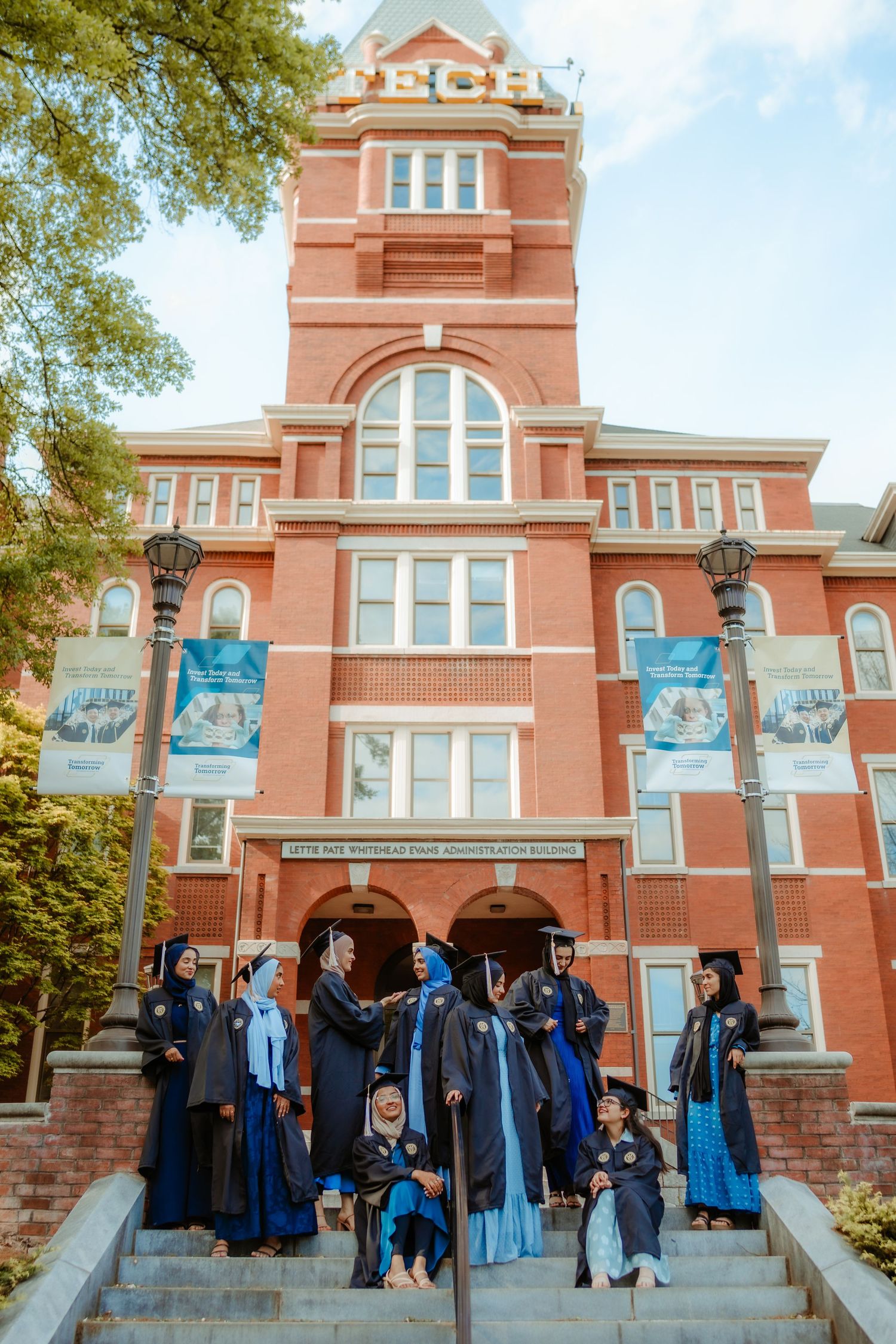 Graduates in blue and black academic regalia congregate on steps of ornate red brick university building.