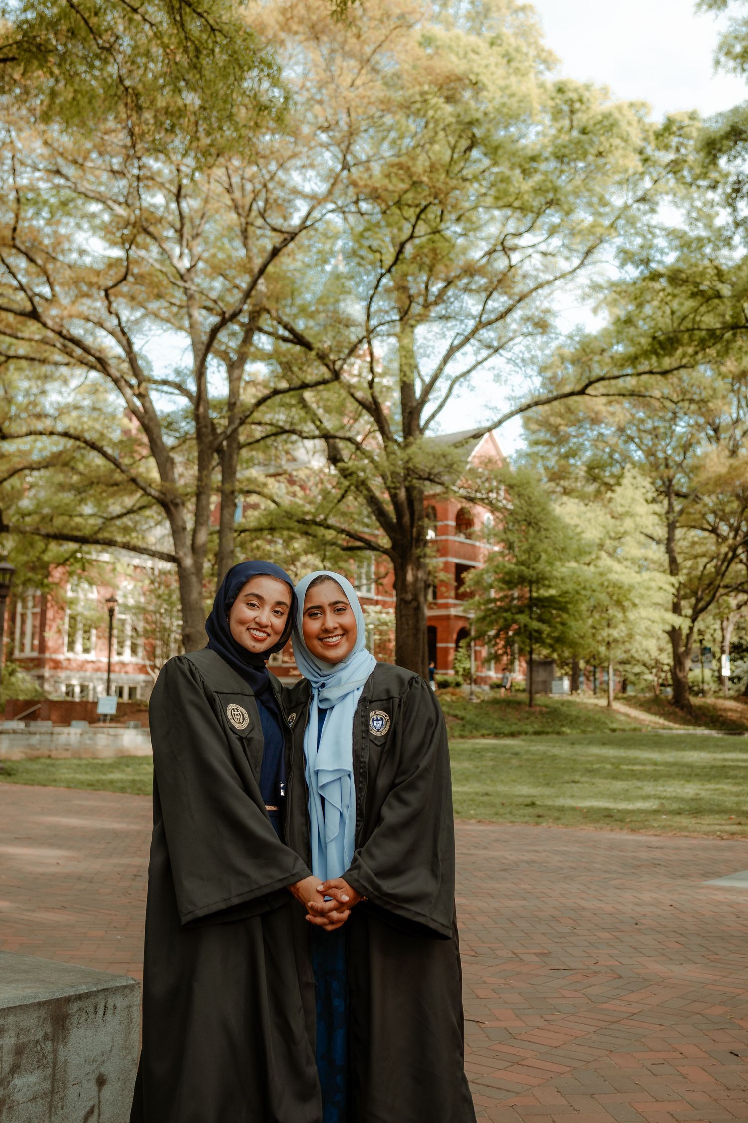 Two graduates in black academic robes celebrate together on brick pathway with university building in background.
