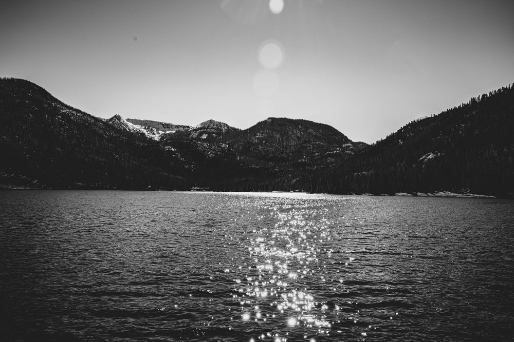 Black and white lake view with sunlight sparkling on water and mountain backdrop.