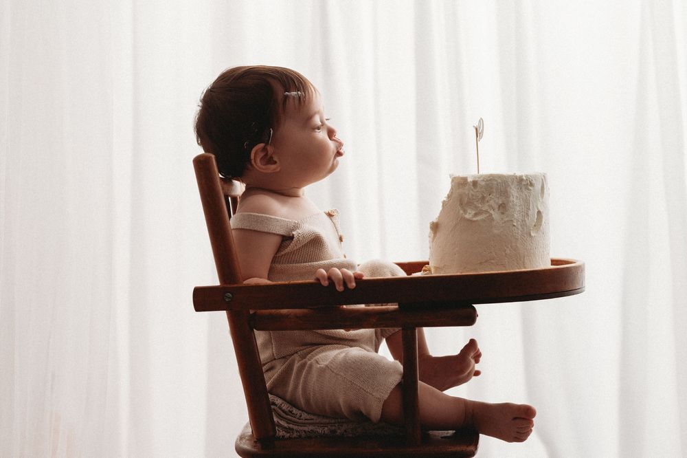 Baby sits in wooden high chair by window while eating from brown tray during mealtime.