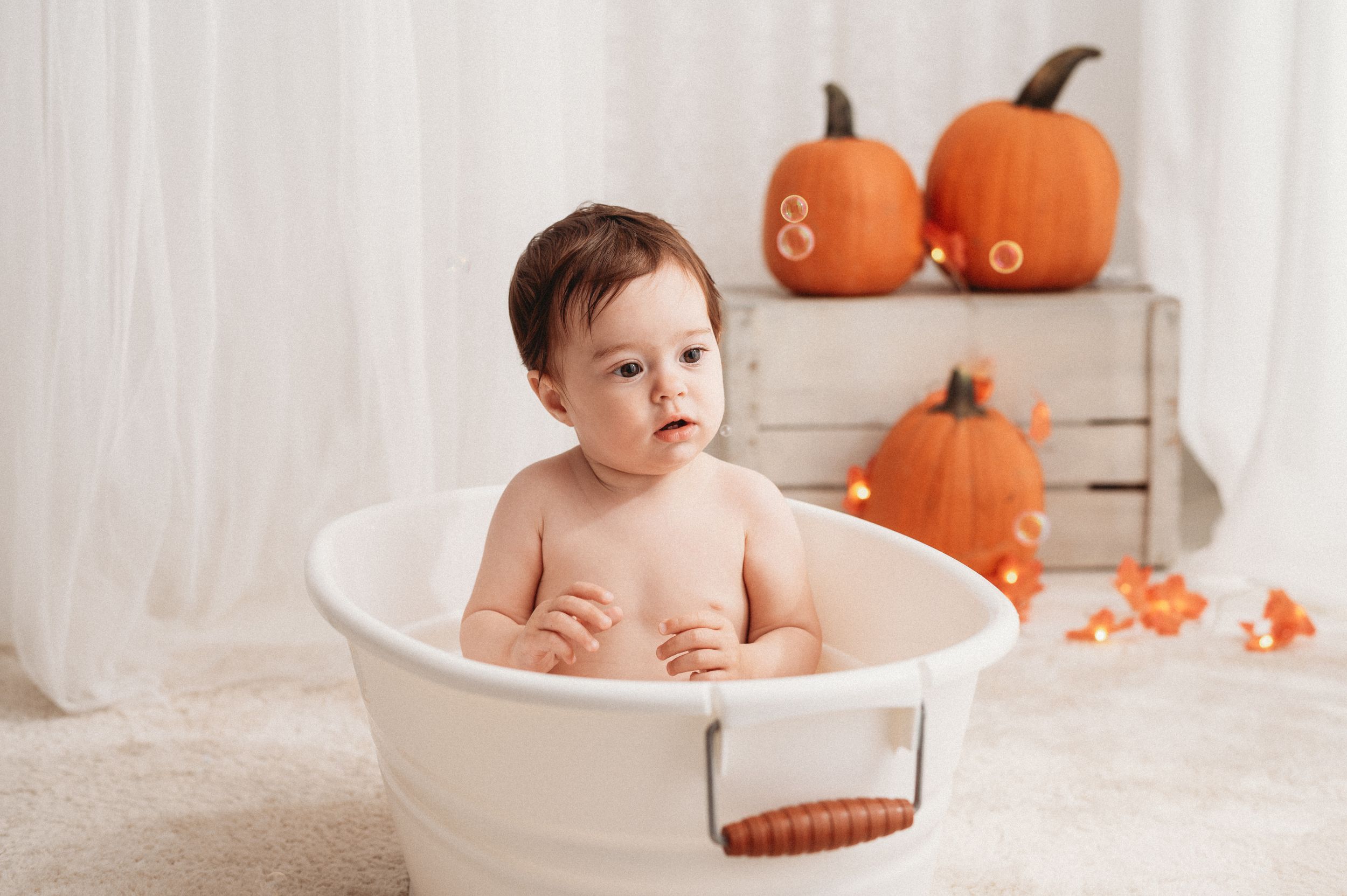 A baby takes a milk bath in a white tub surrounded by small pumpkins for a festive autumn photoshoot.