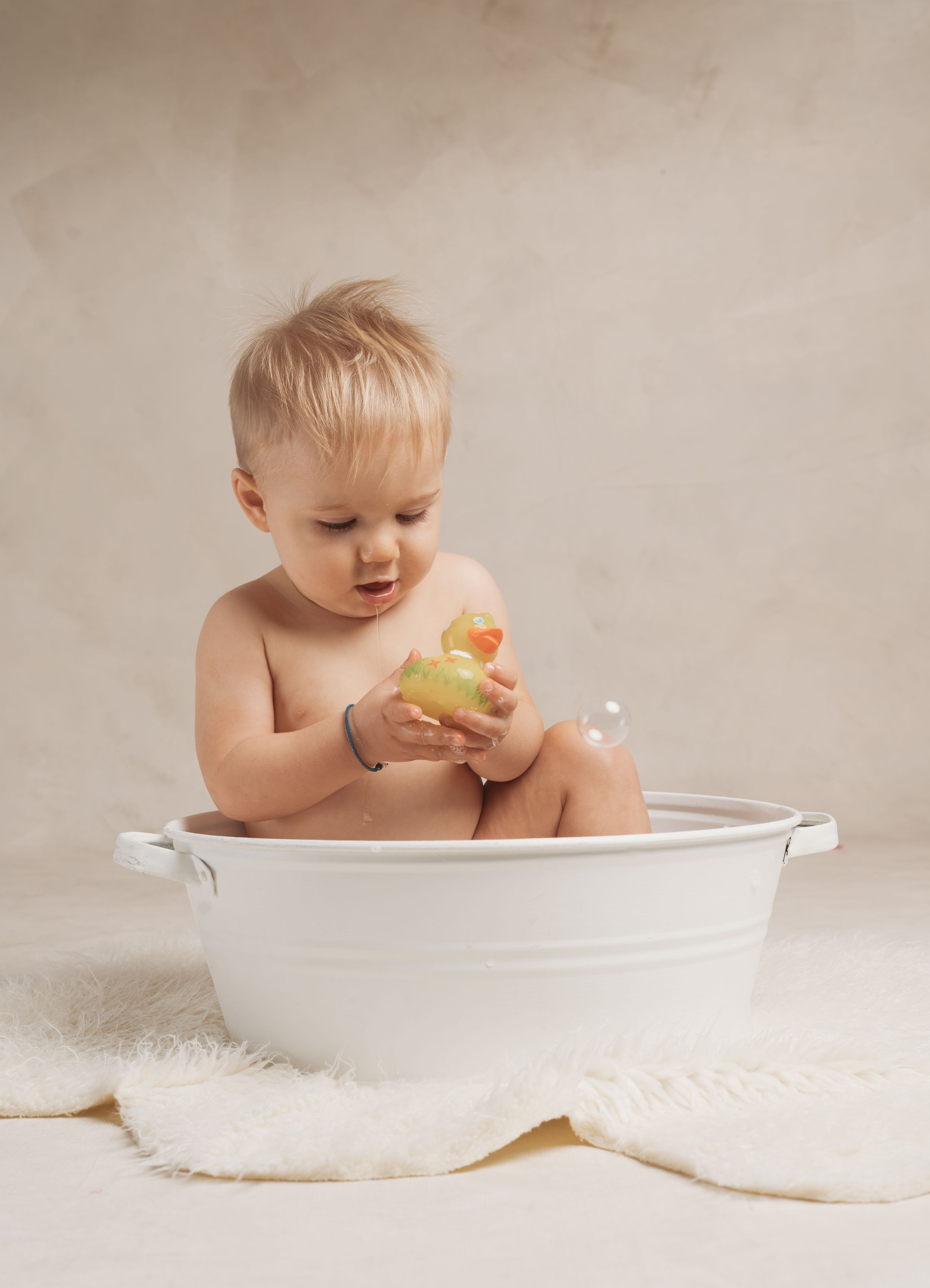 A baby in a white basin taking a bath and playing with a rubber duck against a neutral background.