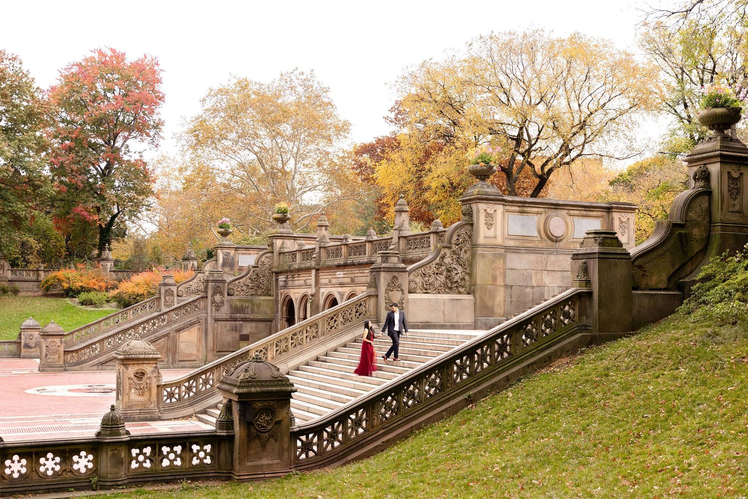 A couple descends ornate stone steps surrounded by autumn foliage in a park setting.