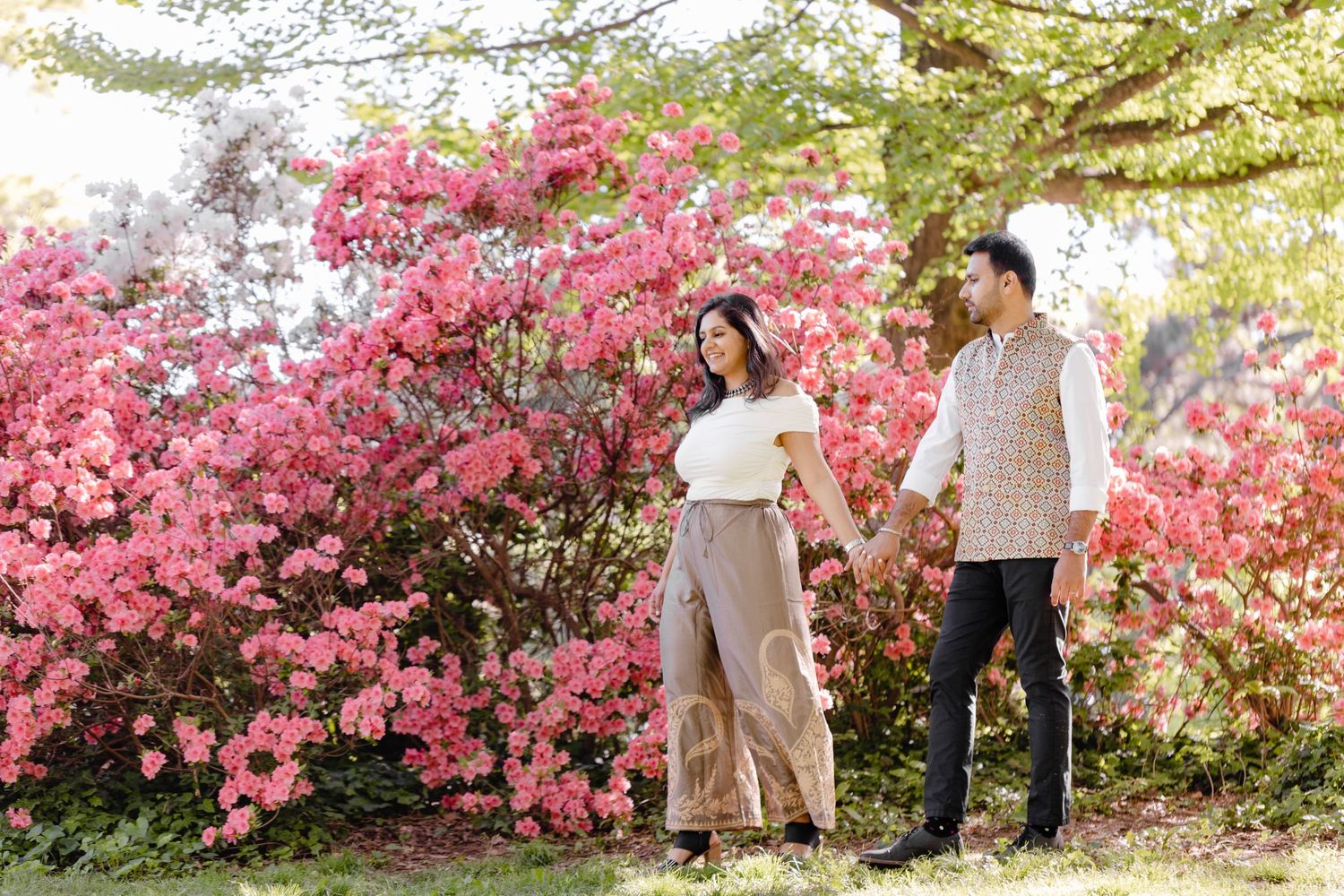 A couple stands together against vibrant pink cherry blossoms in a romantic springtime garden setting.