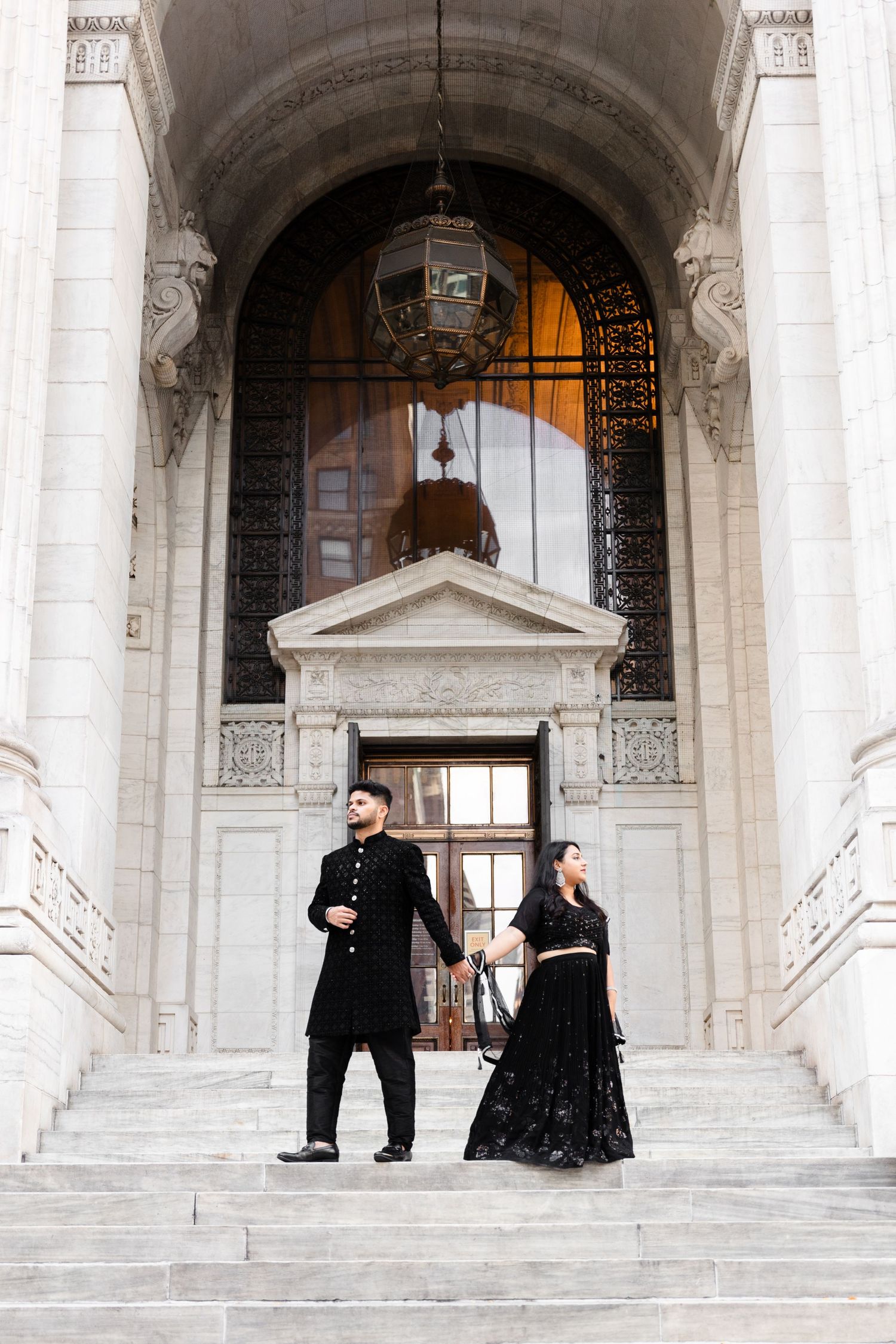 A couple in formal black attire poses dramatically on grand marble steps of a historic building.