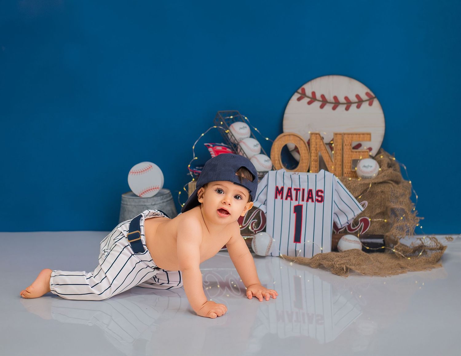 Baby wearing baseball cap lays on blanket with baseball-themed props and decorations for first birthday photo shoot.