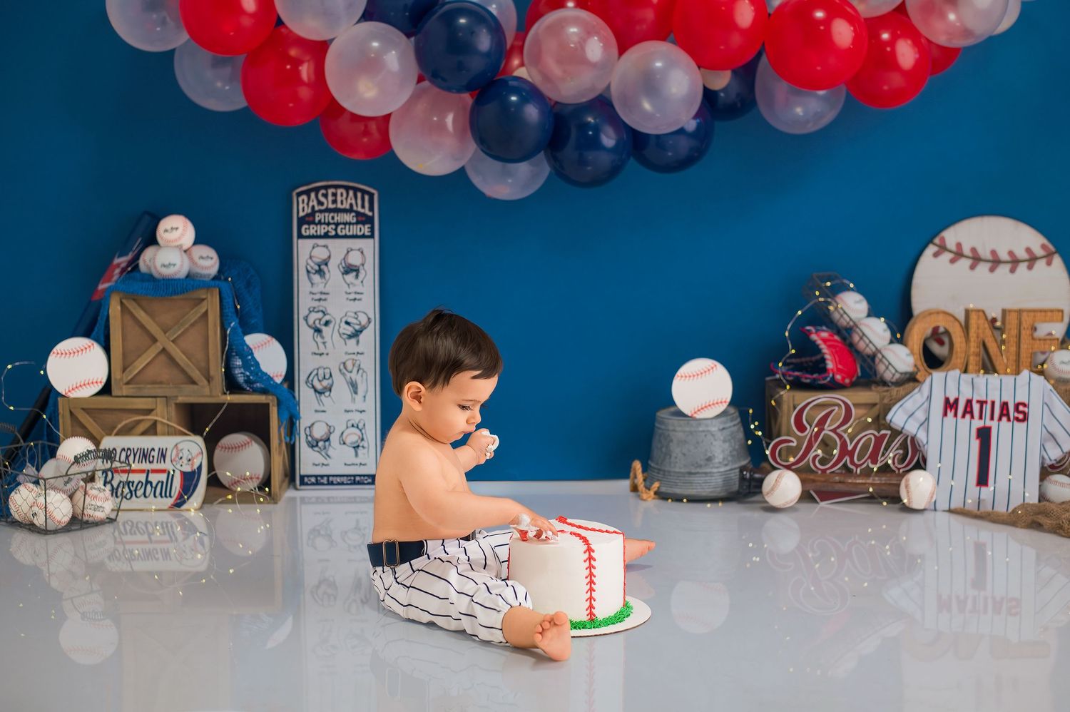 A collection of baseball-themed baby birthday party decorations with white, red and navy blue balloons and banners.
