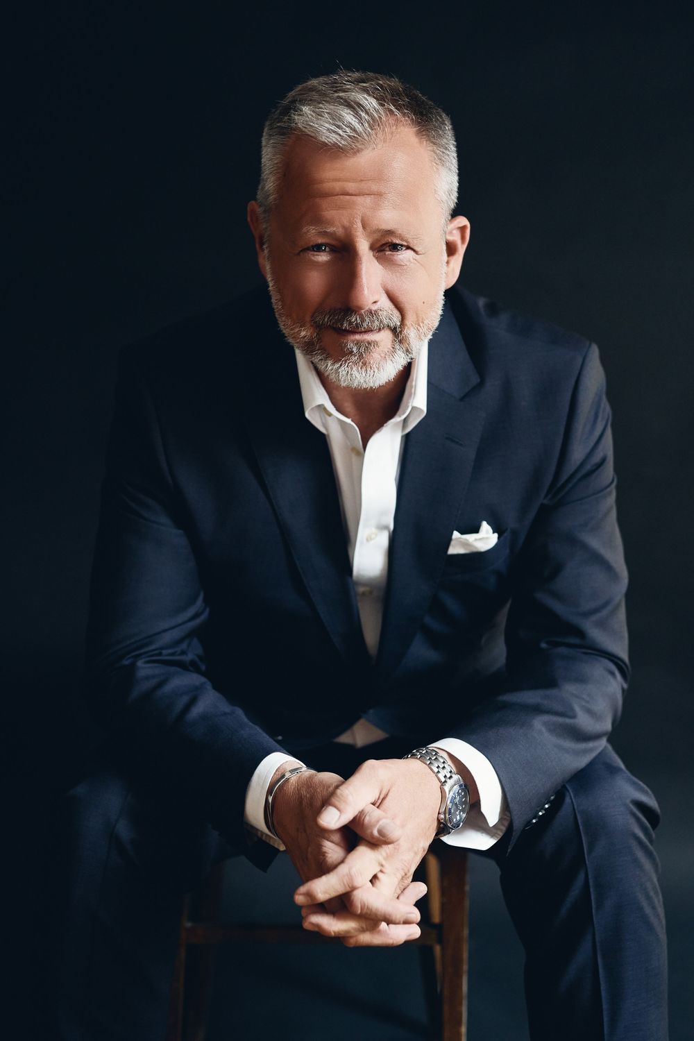 Confident mature businessman with gray hair and beard sitting in navy suit with white pocket square and luxury watch.