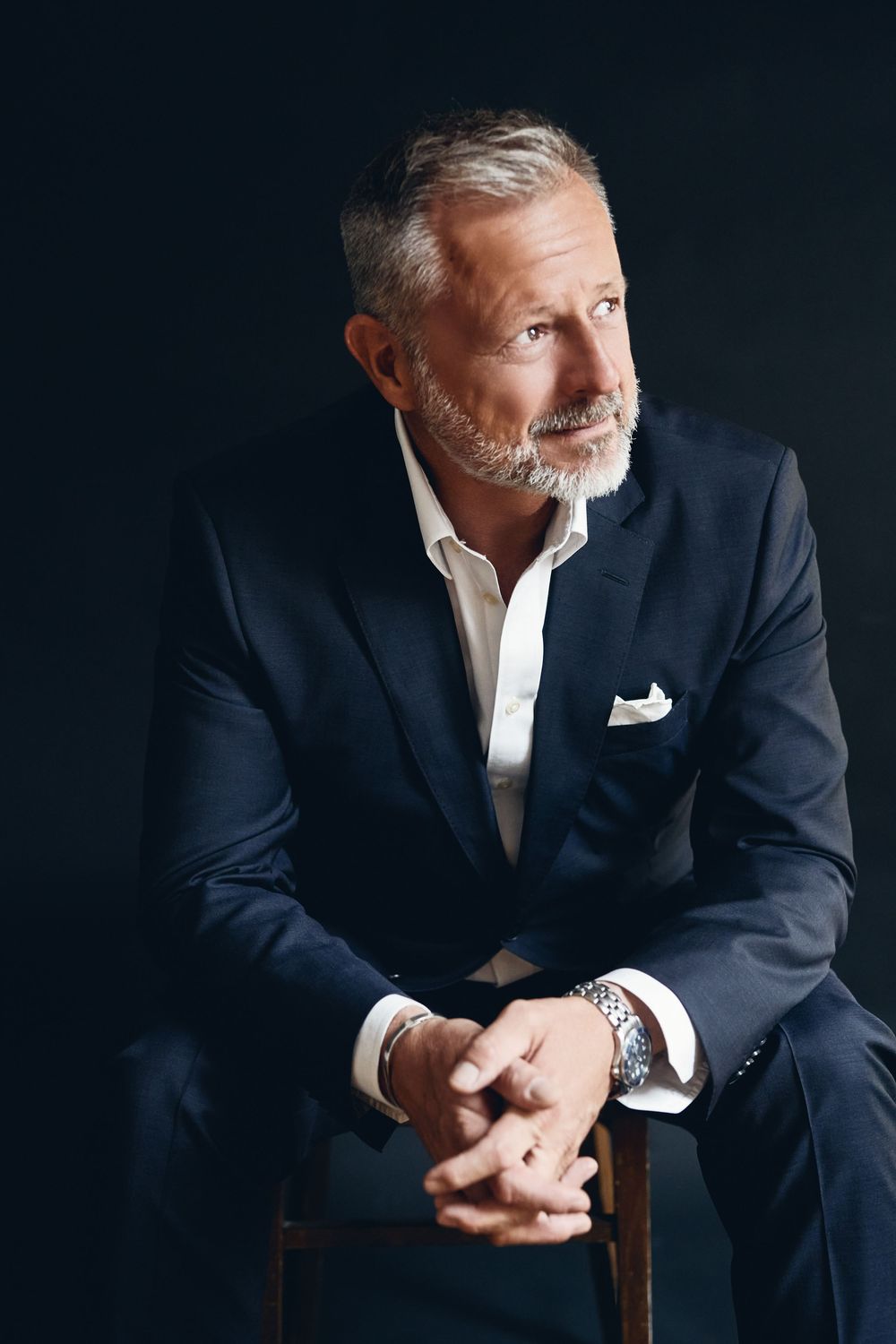Mature businessman with gray beard sits confidently in navy suit with pocket square and watch on dark background.