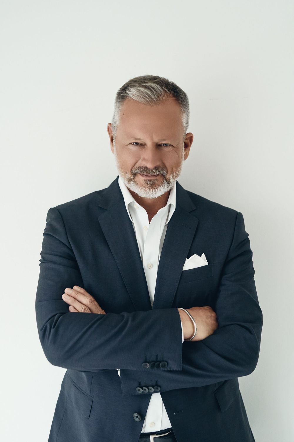 Confident middle-aged businessman with gray hair and beard, wearing dark suit, arms crossed, white background.