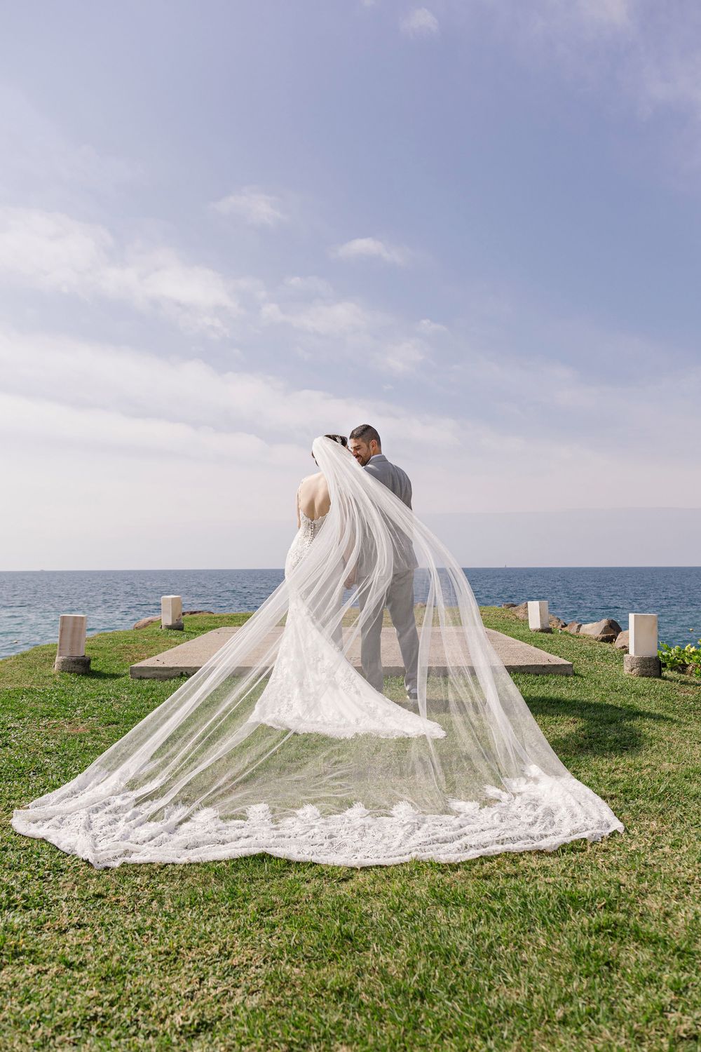 Una pareja romántica se besa bajo un velo blanco vaporoso frente al mar, en un acantilado cubierto de hierba.