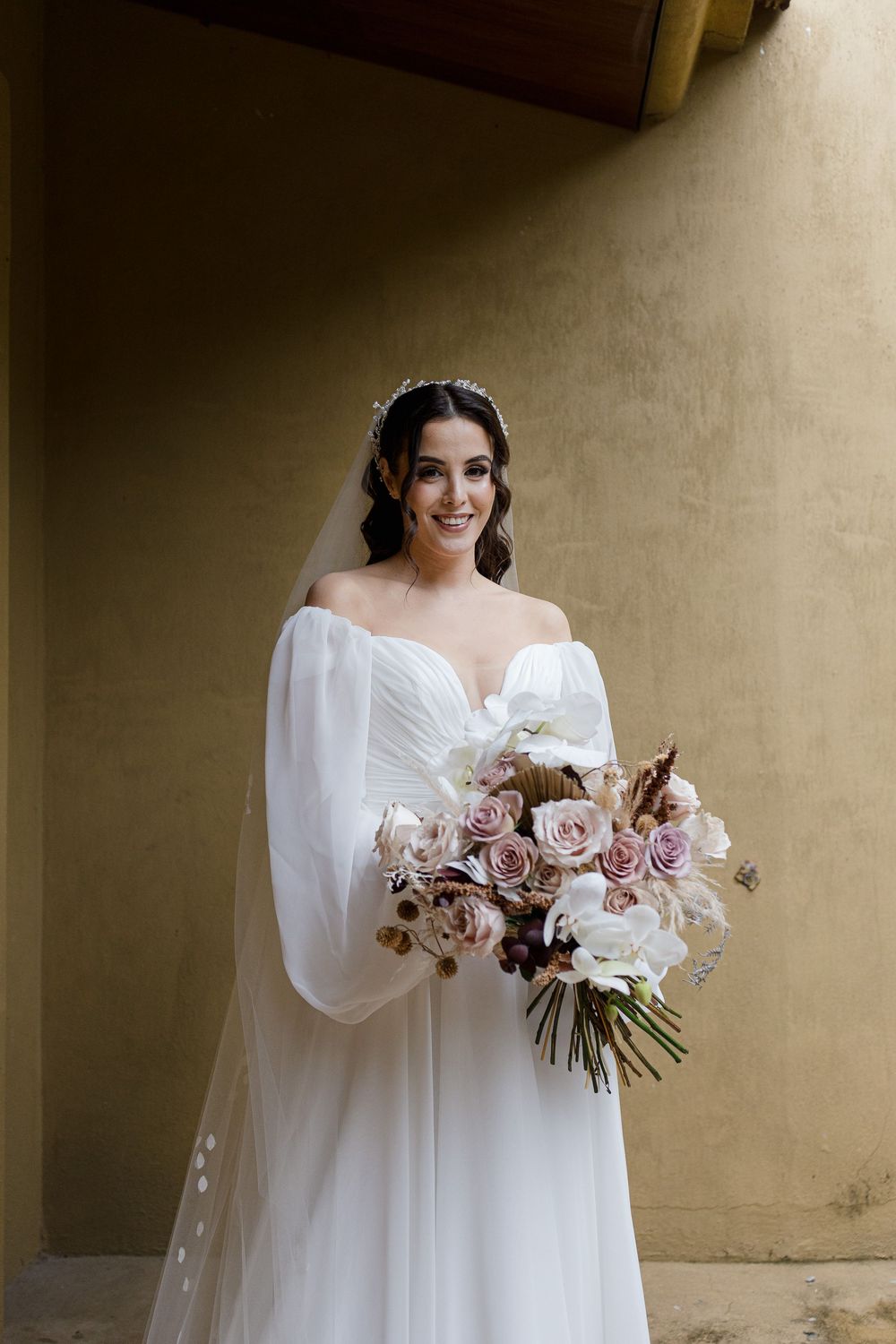 A white wedding dress with flowing sleeves and floral bouquet captured in an elegant indoor setting.