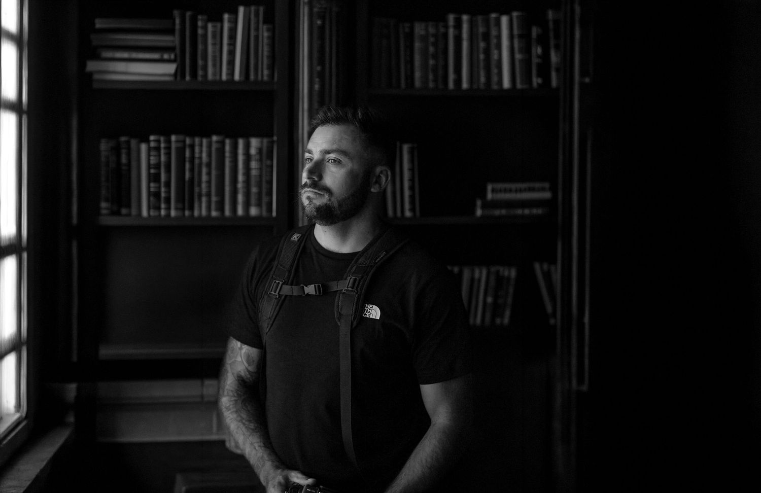 Person in black t-shirt sits thoughtfully in front of bookshelves in a dark room.