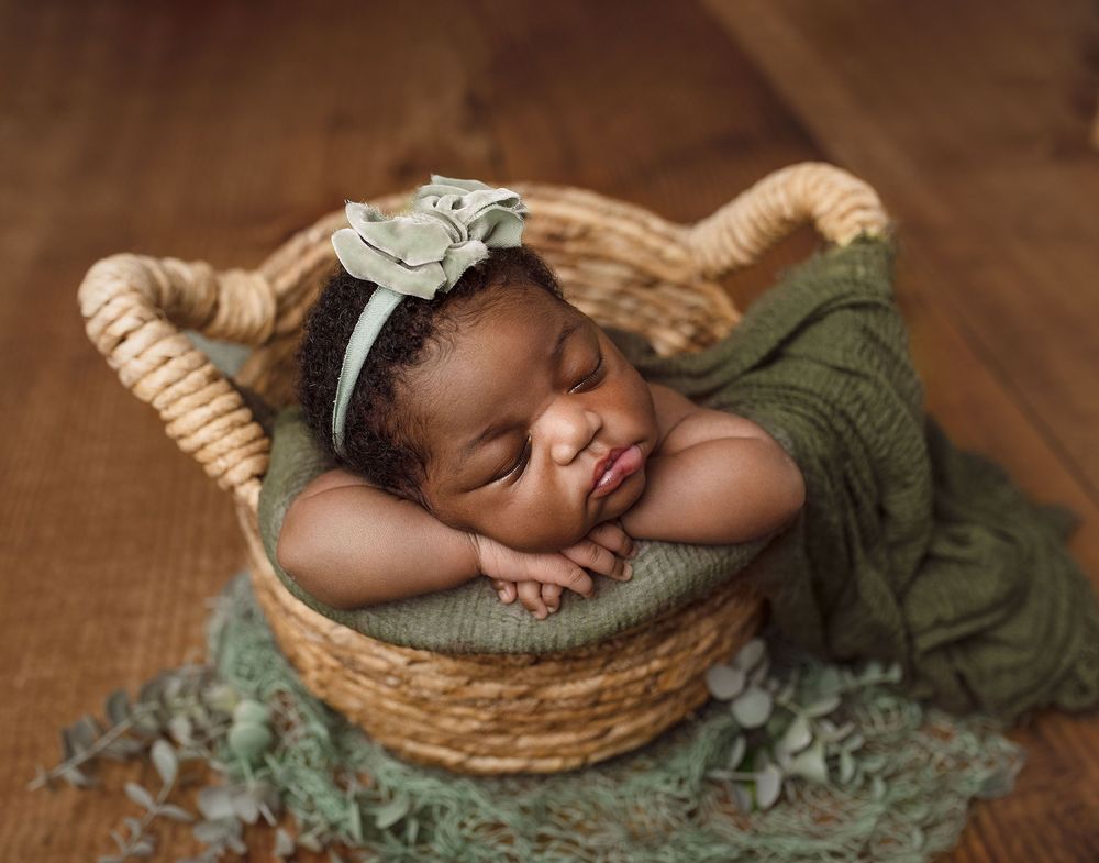 An adorable sleeping newborn wearing an olive knit wrap rests peacefully in a woven basket on wooden flooring.