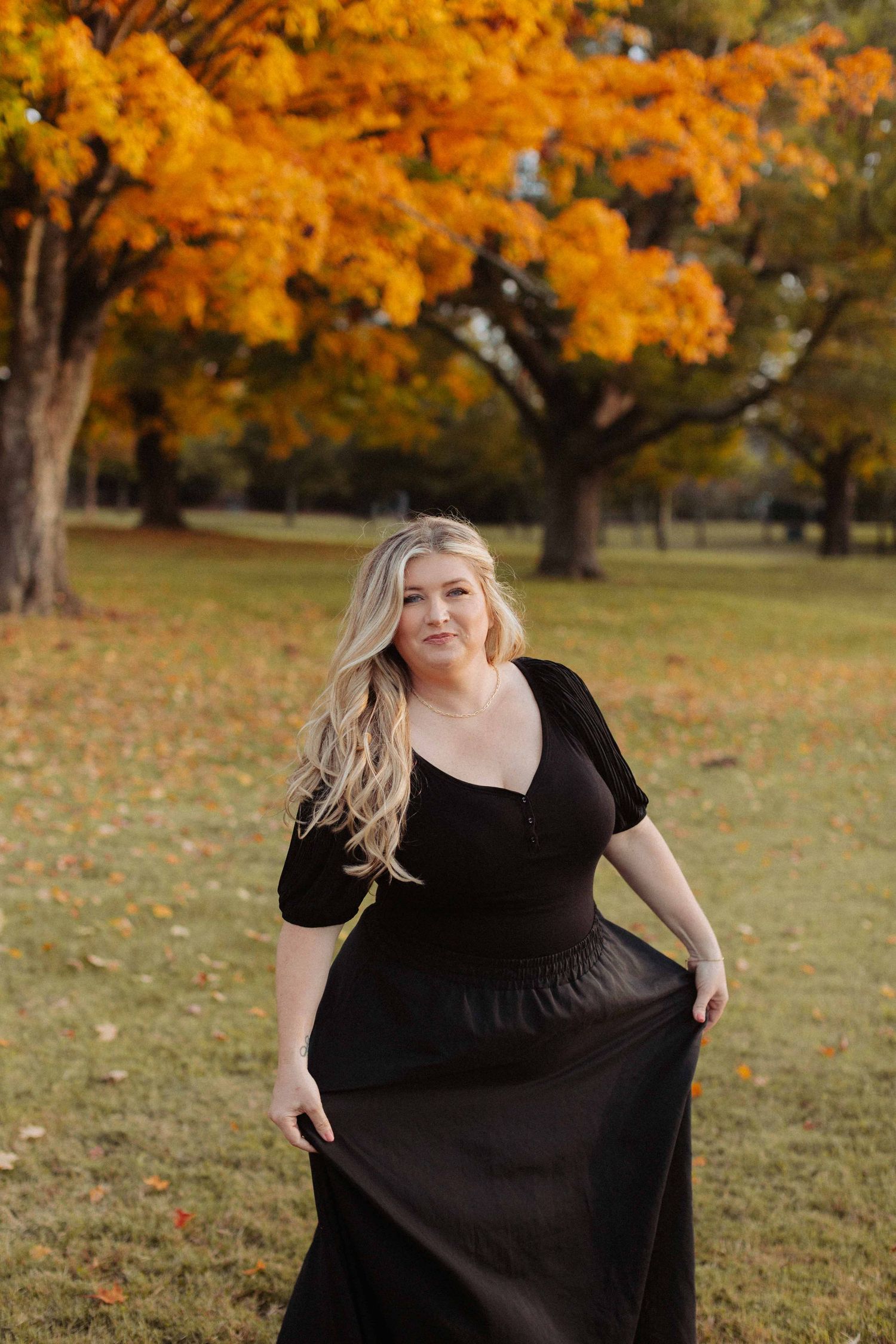 Someone in a black dress poses in a park with vibrant orange autumn trees in the background.