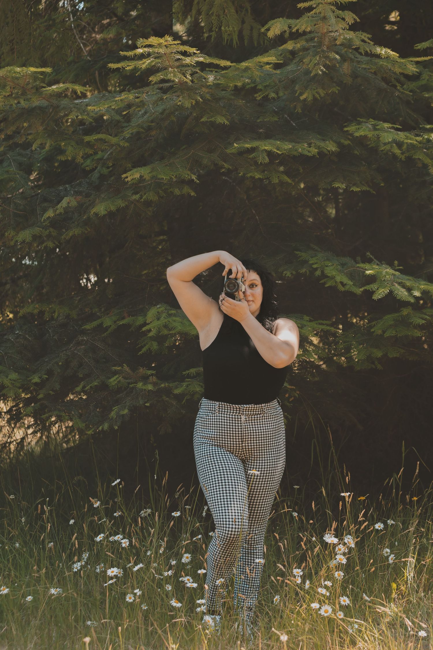 A woman standing in a field of daisys with a tree behind her. She is holding a camera. She is wearing checkered pants and a black shirt.