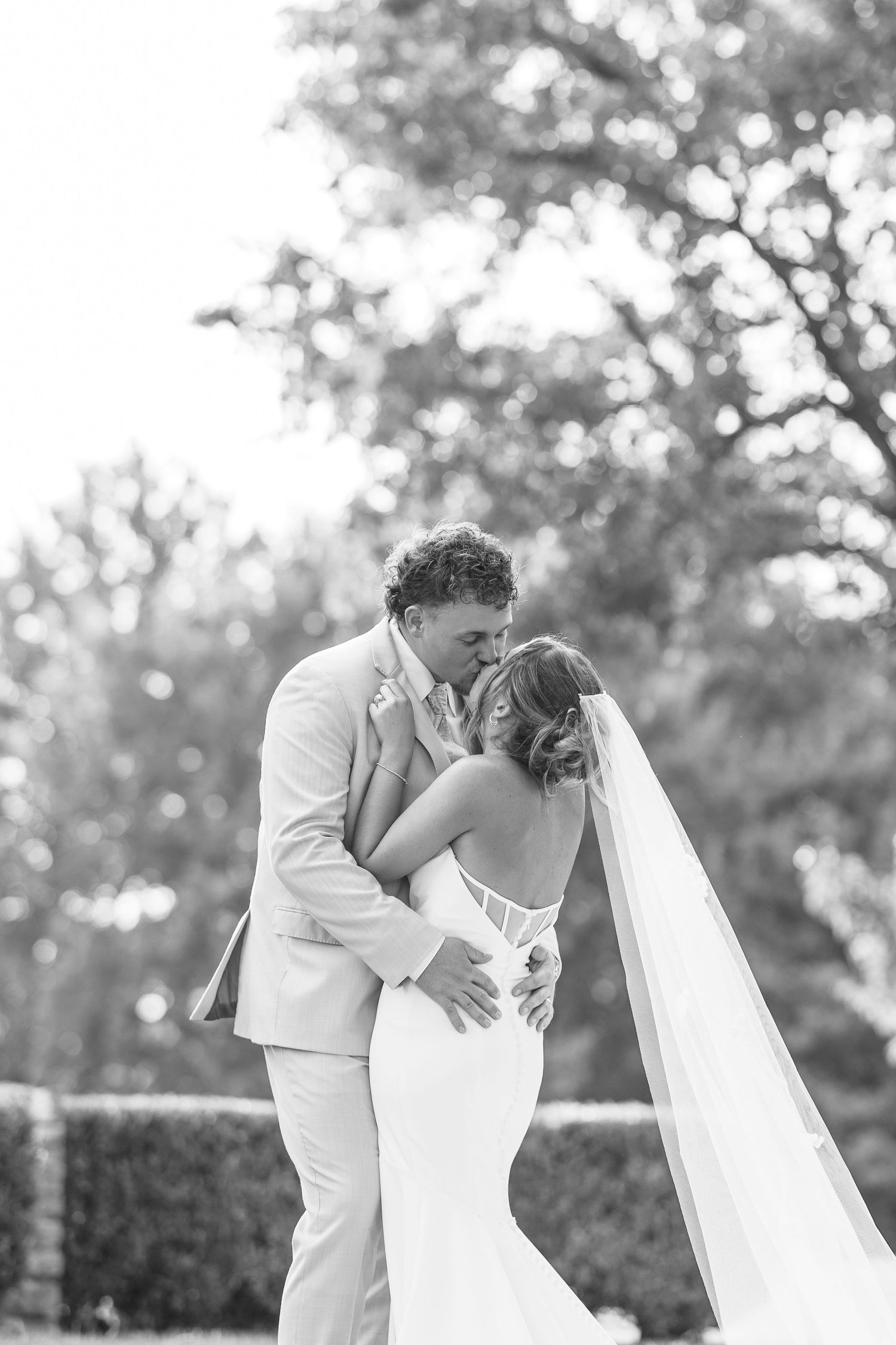 A romantic black and white wedding moment captured outdoors with a bride in a fitted dress and flowing veil.