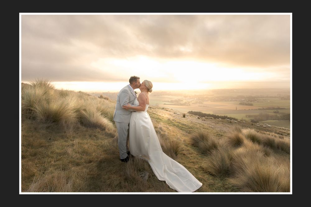 Bride and groom kissing on a hilltop at golden sunset, with sweeping countryside views and tall grass.